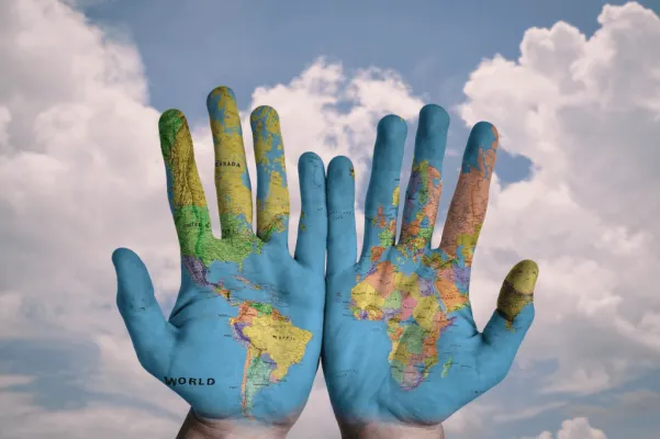 Two open hands painted with a world map, the Americas on one and Europe and Africa on the other, held up against a cloudy blue sky