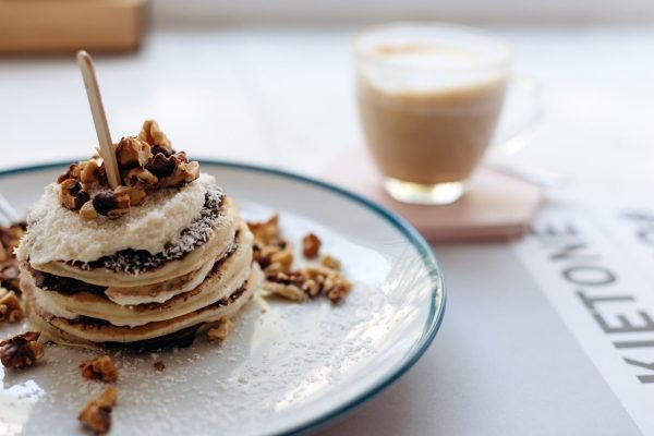 A stack of pancakes topped with nuts, coconut, and chocolate, served with a cup of coffee.