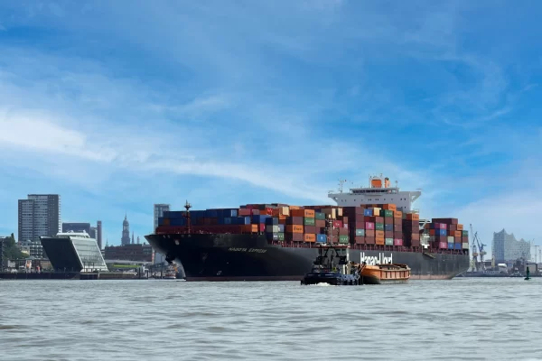 Large container ship carrying cargo containers through a busy commercial port, representing international trade, logistics, and global supply chain operations.